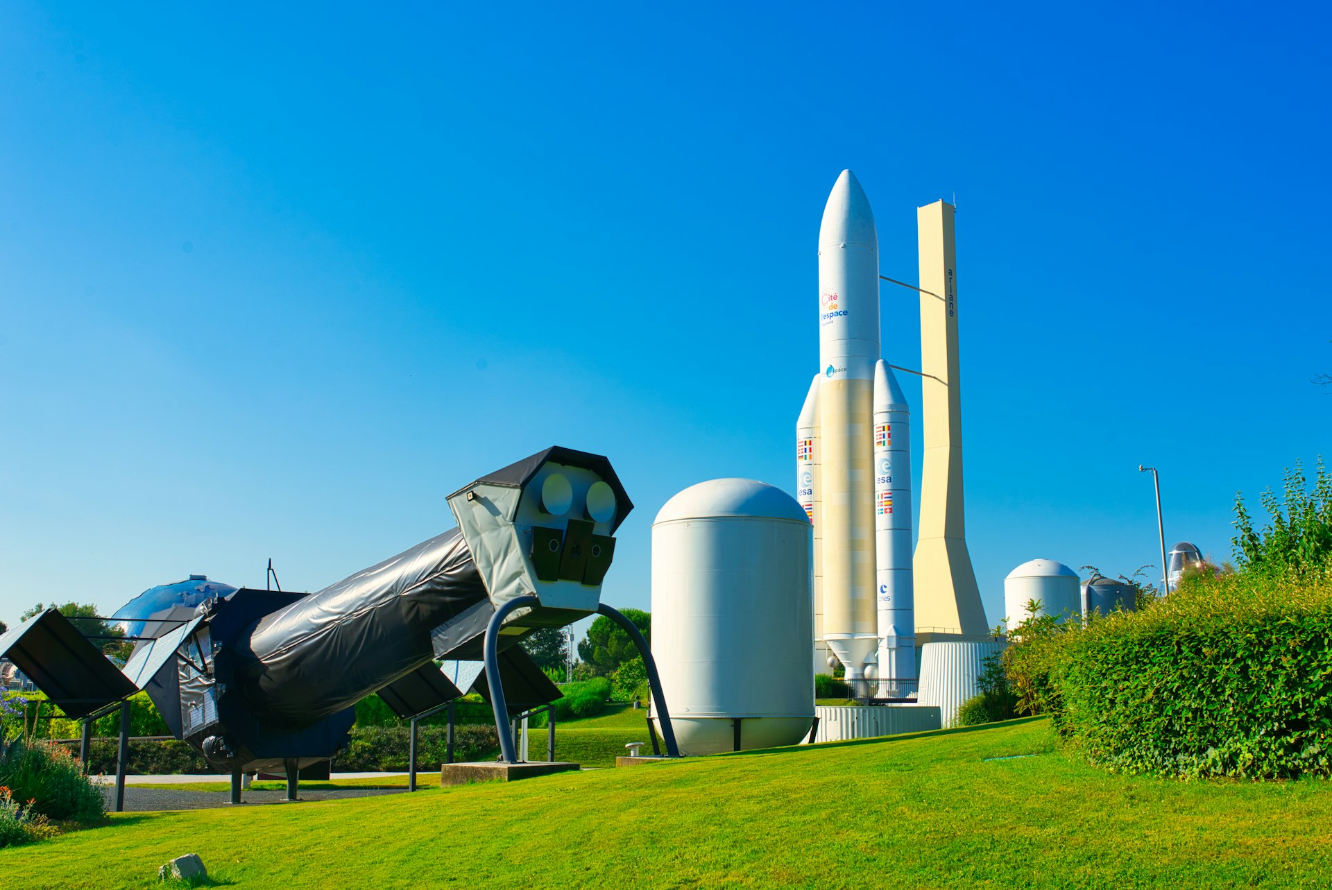 A huge white tubular space rocket stands on its end pointing into the sky. Nearby is a large black satellite