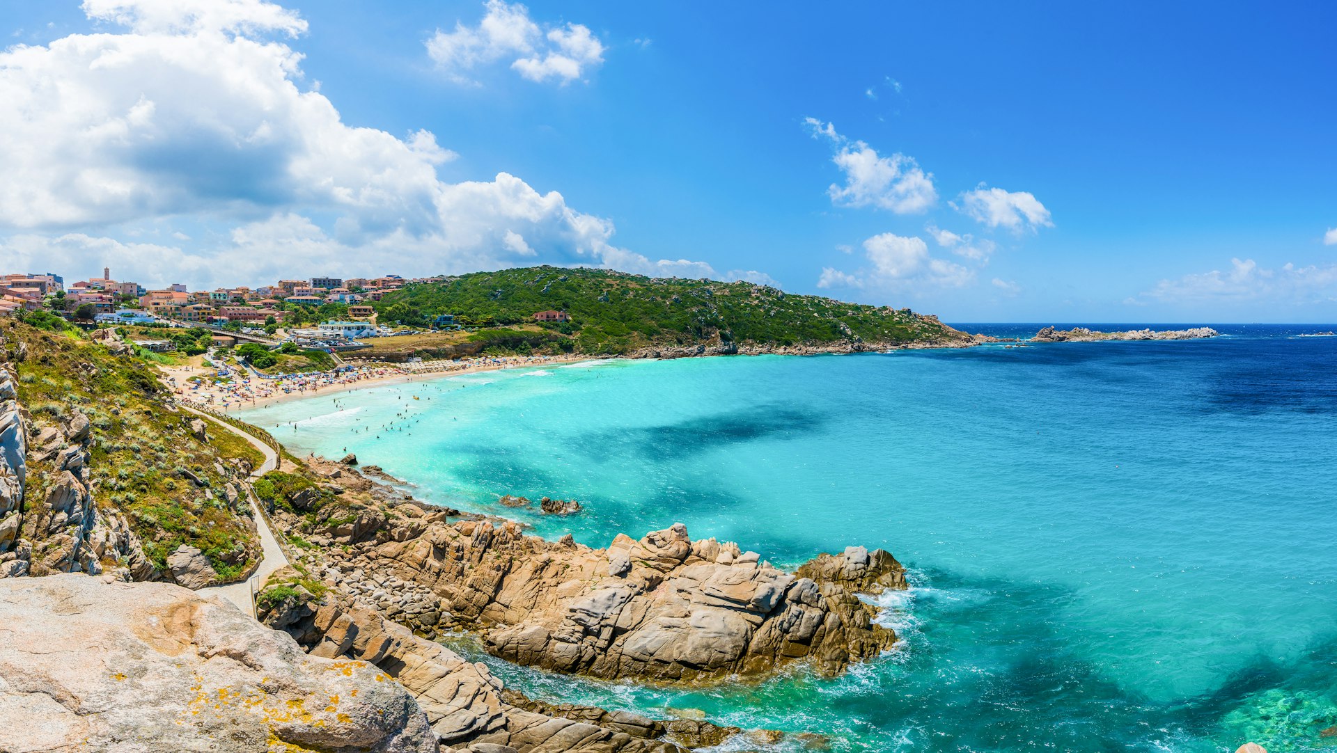A wide angled view of a large cove with shallow pale blue waters and a golden stretch of beach which is full of people.
