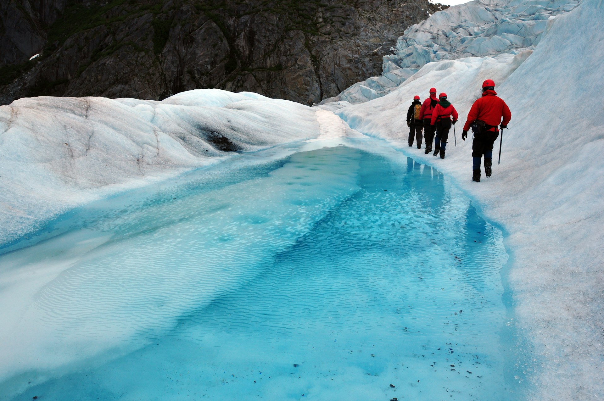 A group of hikers walk across a glacier near a pool of glacial meltwater