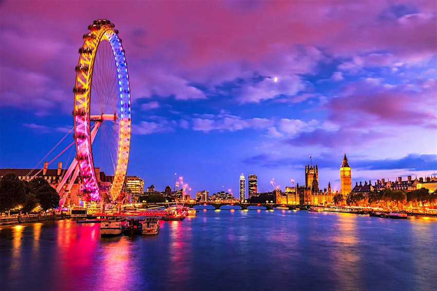 London Eye and the Palace of Westminster A night time shot of a river with a large Ferris wheel lit up in pink, yellow and blue to the left, and a huge neo-Gothic building to the right