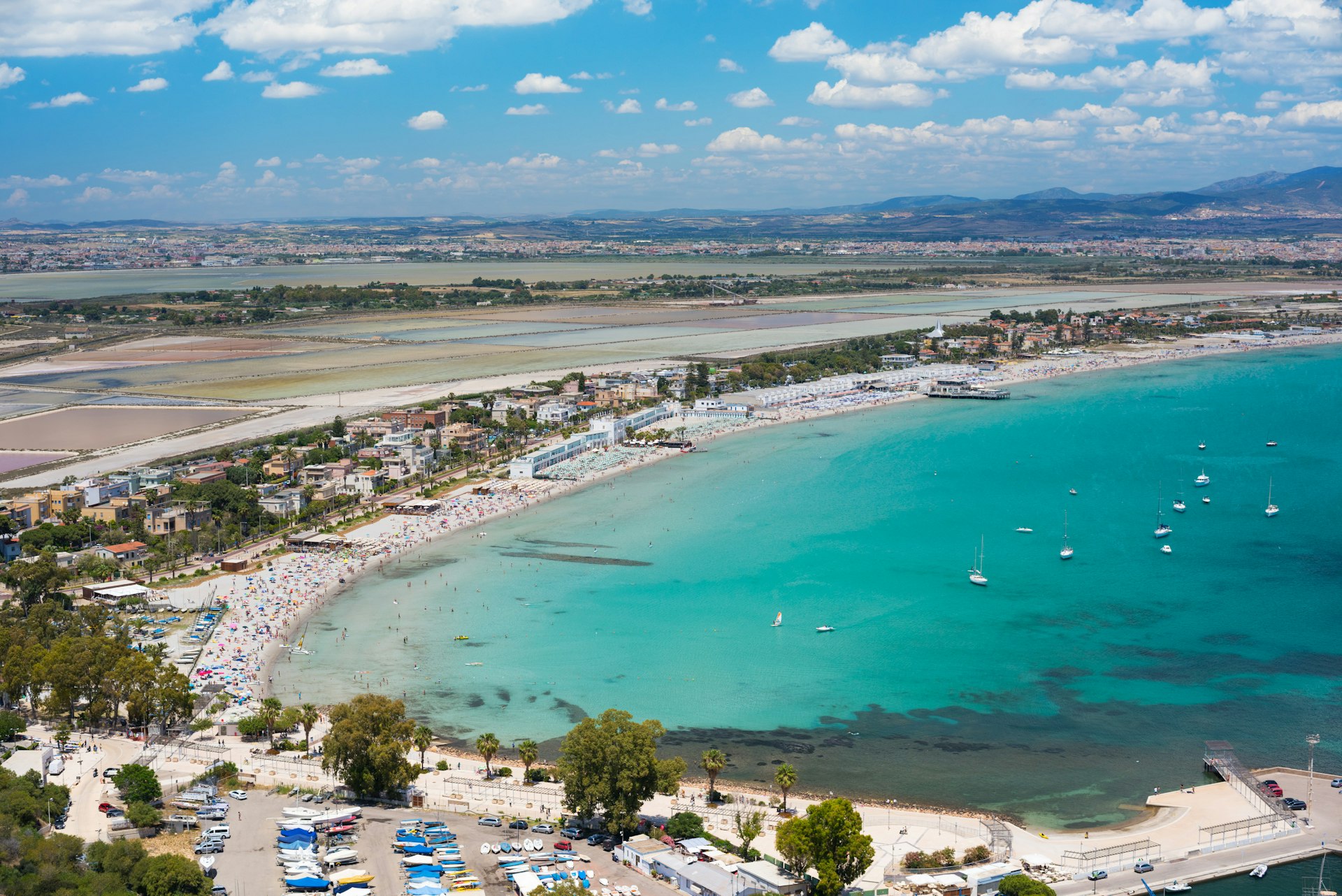A very high-angled view of Poetto Beach, a very large sandy bay with clear blue water. The beach has many people sunbathing on it.