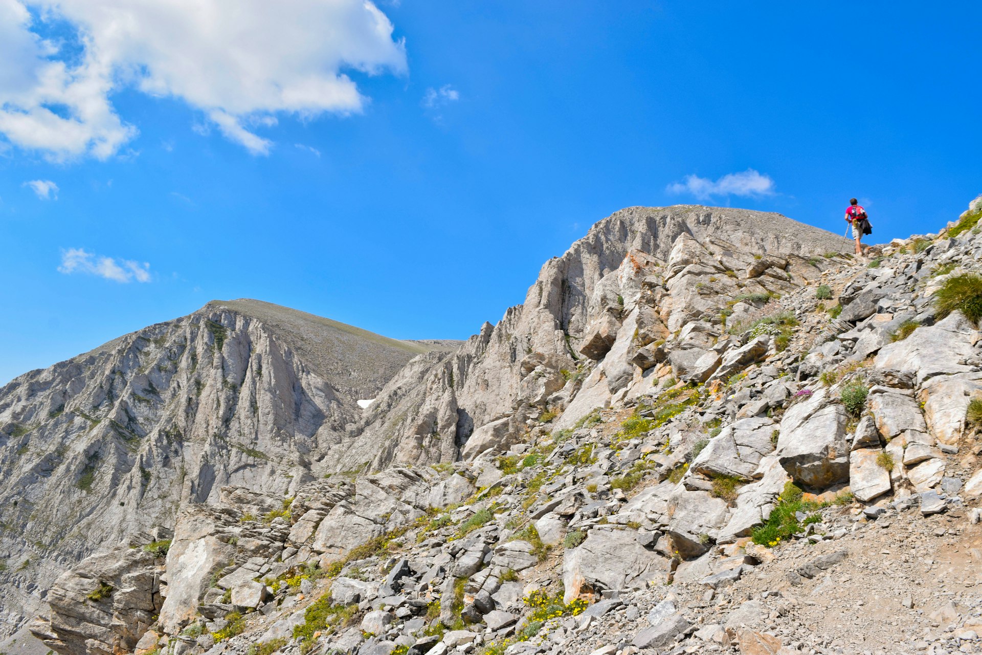 A hiker walks up a steep, rocky path to the peak of Mt Olympus. The mountain peak is bare and rocky and a blue sky is visible above.