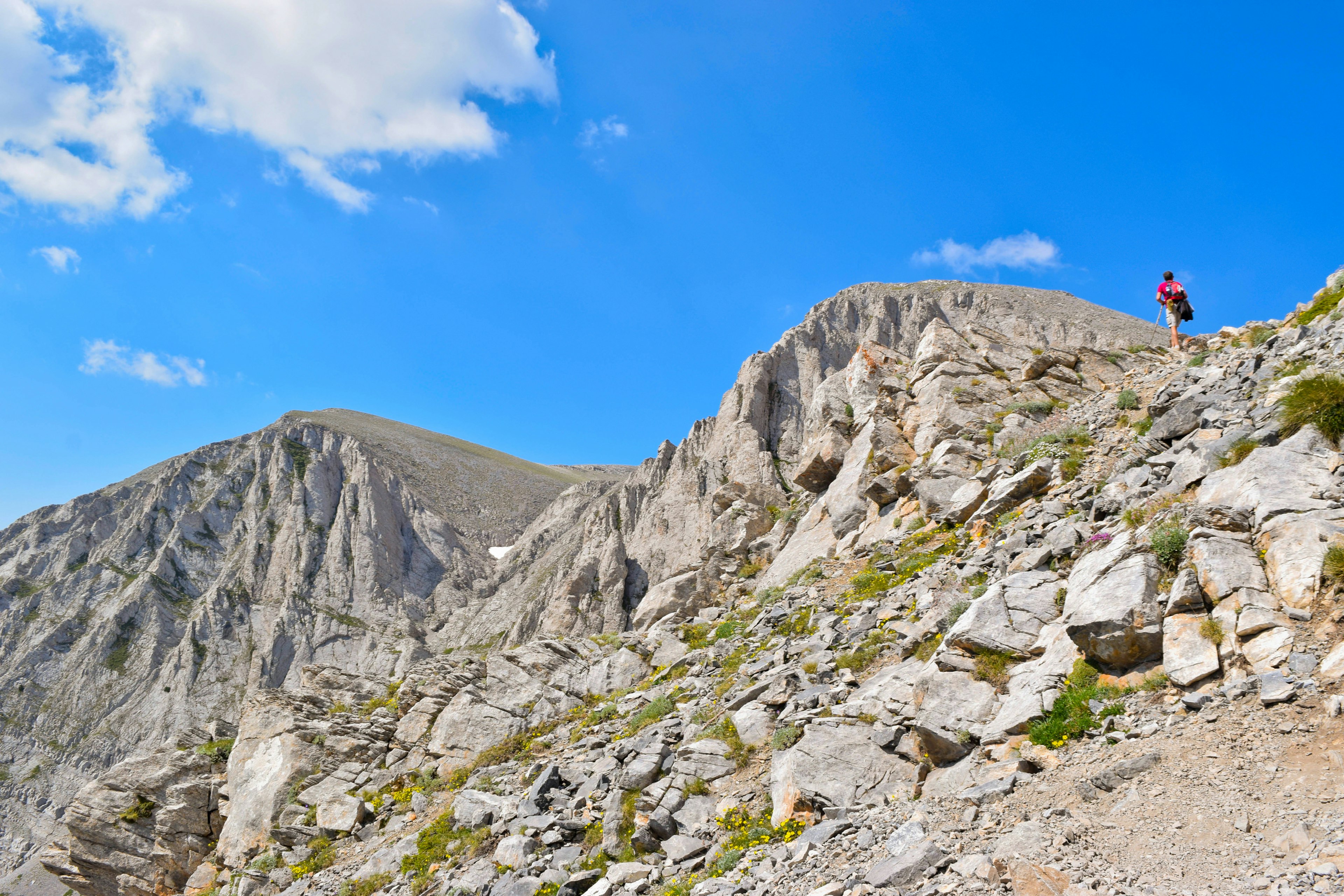 A hiker walks up a steep, rocky path to the peak of Mt Olympus. The mountain peak is bare and rocky and a blue sky is visible above.