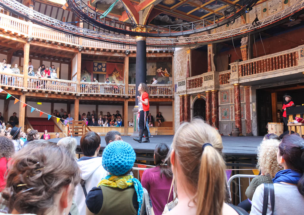 Shakespeare's Globe A crowd of people standing below a stage in a wooden theater. A person on the stage addresses the crowd
