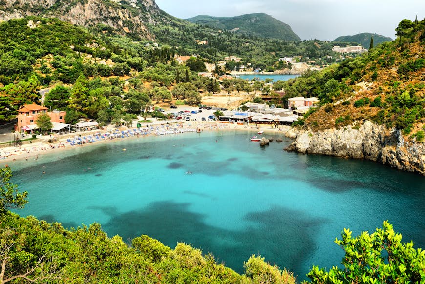 A view of Paleokastritsa Bay, Corfu. The water is an inviting turquoise blue, while a number of tourists lounge on a curving strip of white sand. Beyond them are green hills.