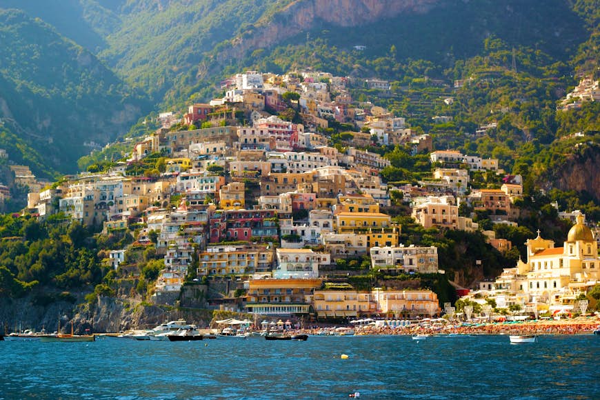 Färgglada hus täcker en brant kustsluttning, som utgör en del av staden Positano på Amalfikusten, Italien.  Bilden är tagen från havet och båtar ligger förtöjda i vattnet nära stranden.