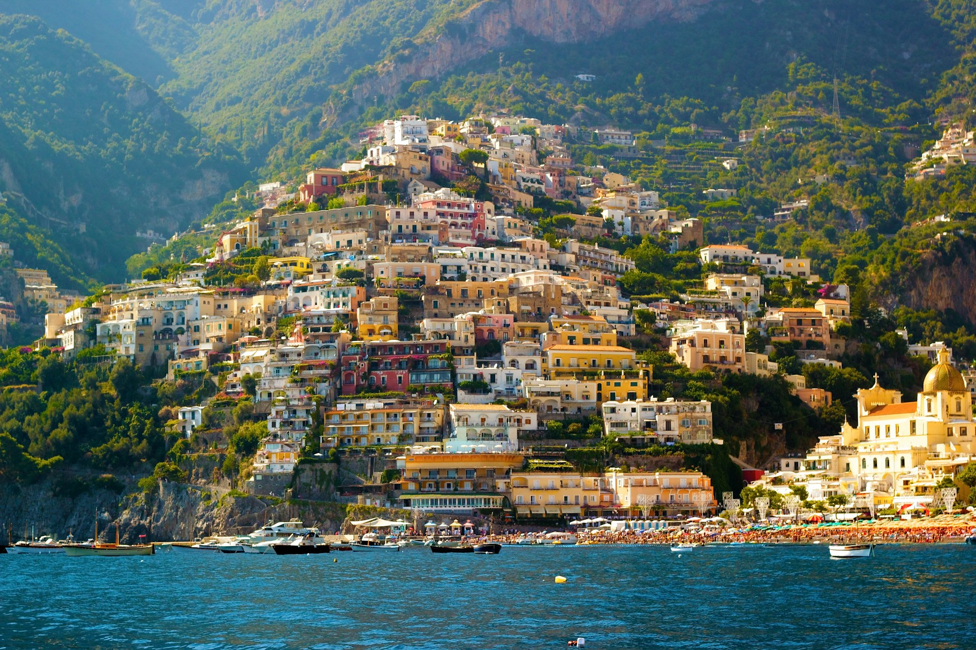 Colourful houses cover a steep coastal hillside, making up part of the town of Positano on the Amalfi Coast, Italy. The photo is taken from the sea and boats are docked in the water near the shore.