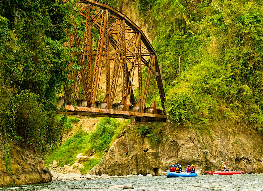 Rafters paddling under an old railroad bridge with kids swinging from a rock, in the Lower Pacuare River.