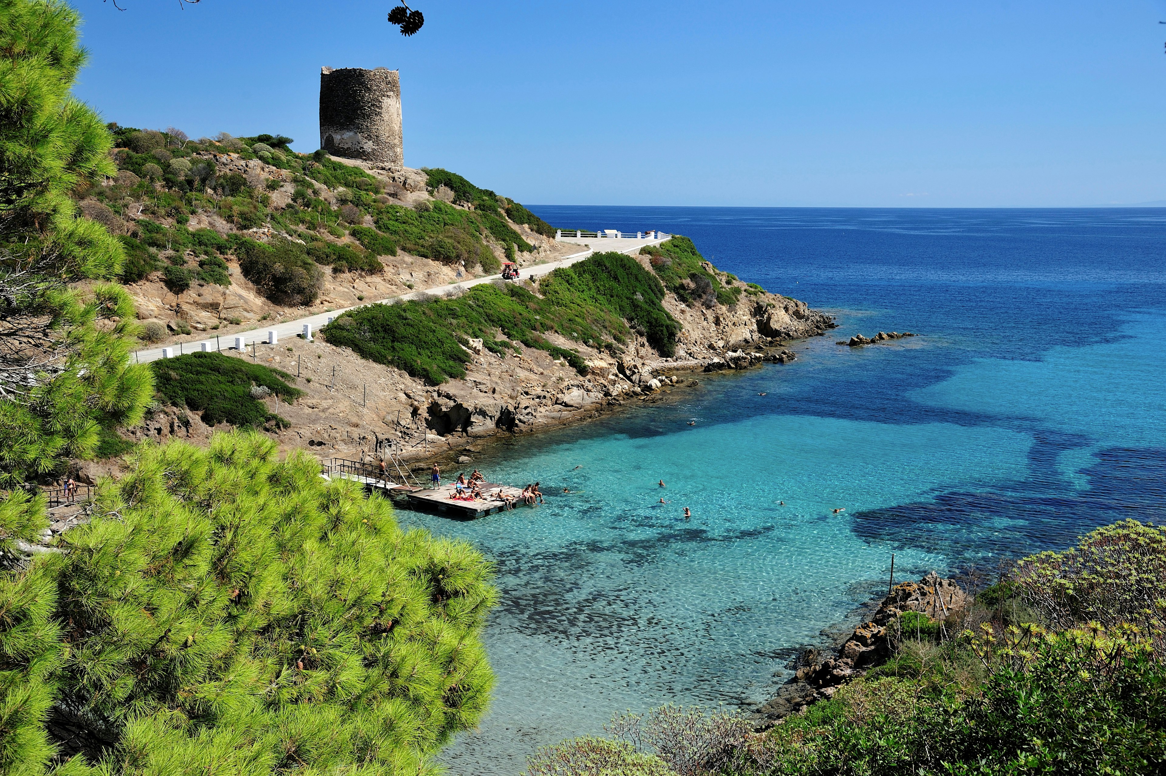 Cala d'Oliva bay on Asinara Island. The sheltered bay boasts shallow blue water, and a handful of people lounge on the rocks next to it. In the background a fortification tower of some kind stands atop a hill.