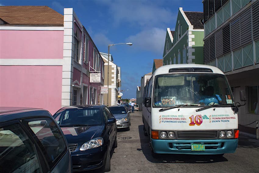 Bahamas Bus drives down a street, there are a row of park cars on the left side of the street