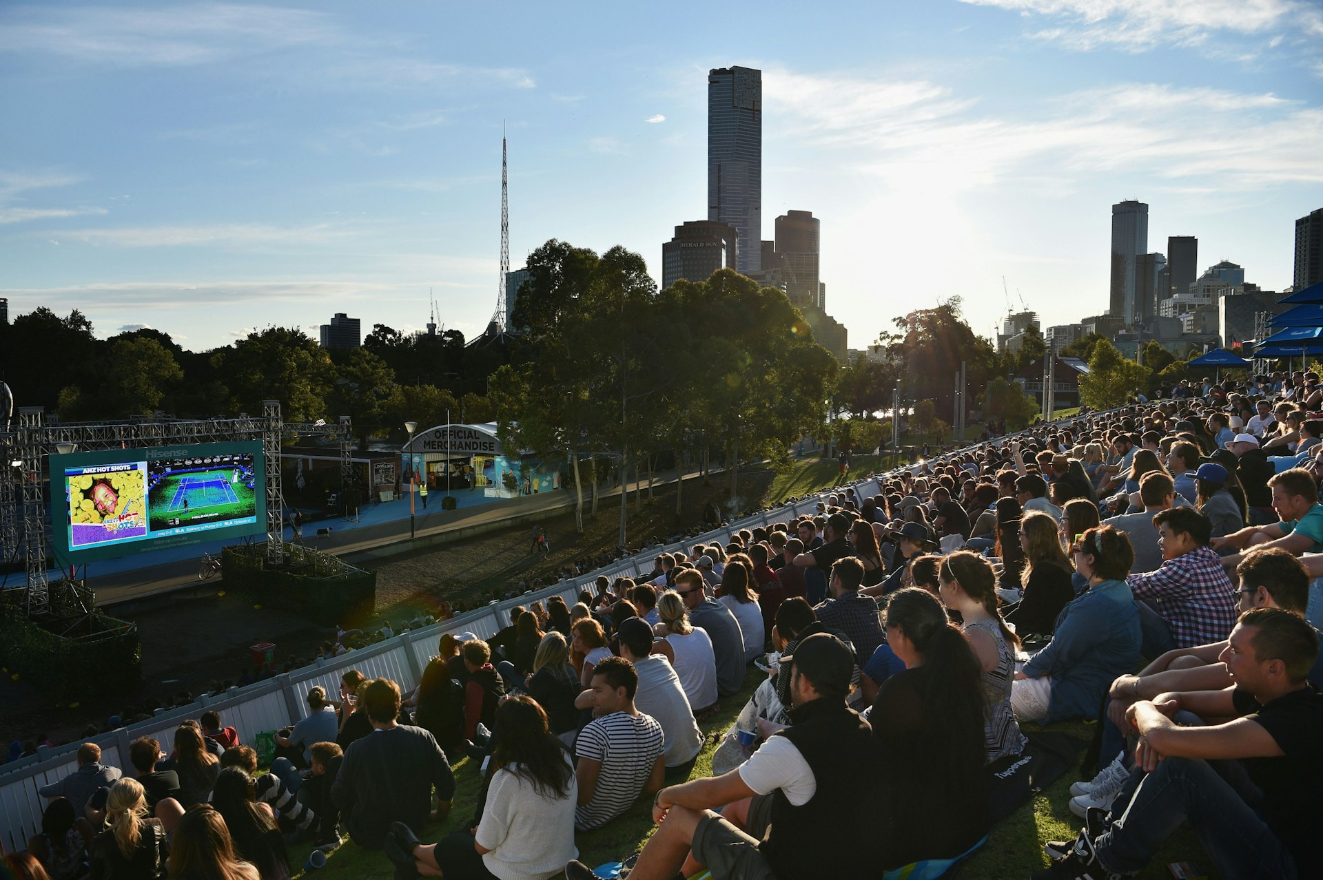 Crowds of people sit on a sloping grassy hill in Birrarung Marr watching the Men's Singles Final match of the Australian Open on a large outdoor screen.