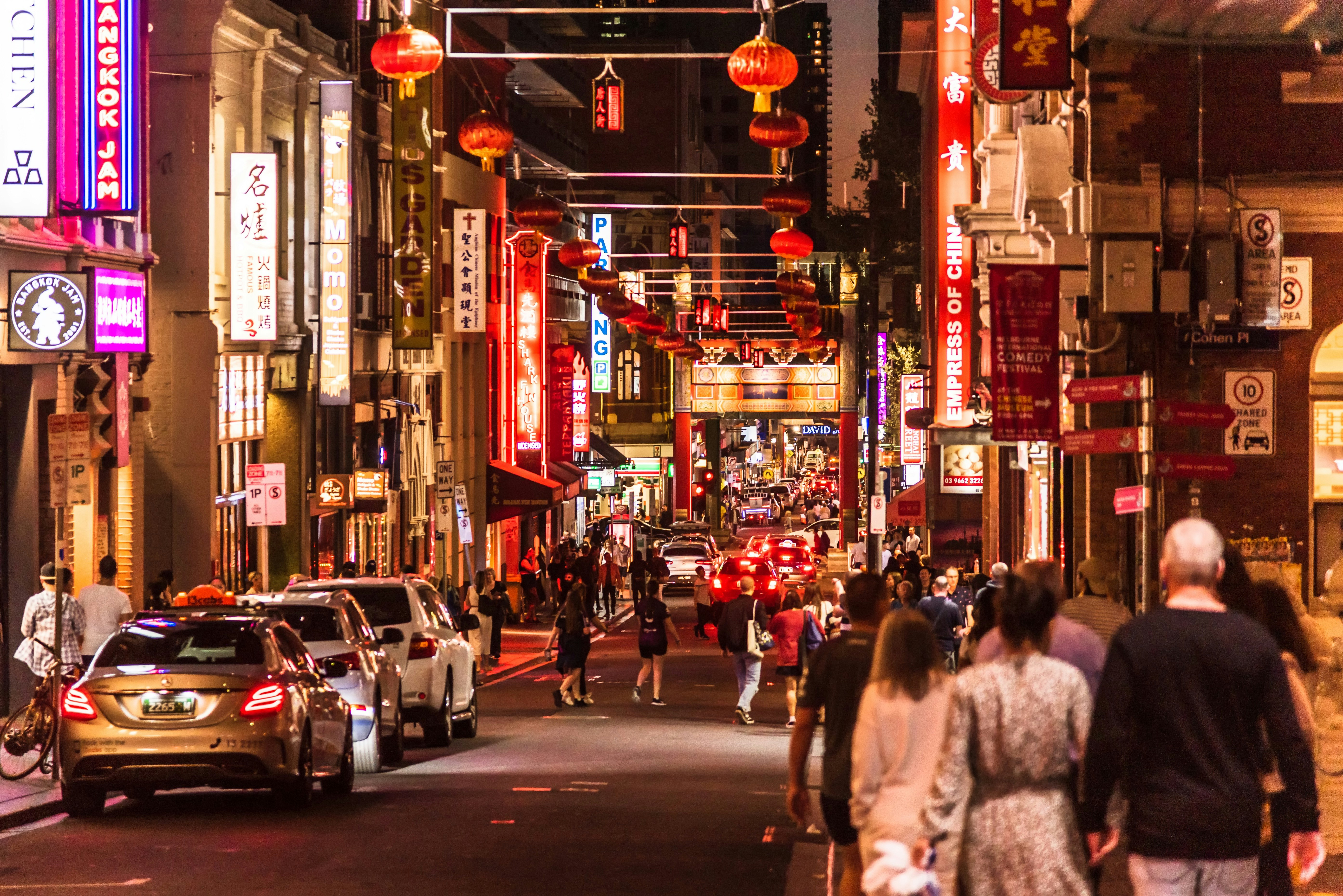 An evening view of people walking through the streets of Chinatown in Melbourne. Red lanterns dangle from buildings and arches above the street.