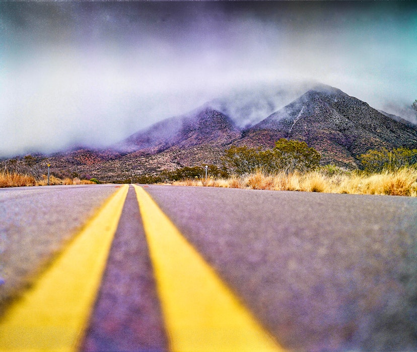 Low angle view near the yellow stripes of the road that heads towards the Franklin Mountains mountains covered by clouds, creating a misty atmosphere. Shrubs and grasses of a vivid golden yellow color grow along side the road. El Paso, Texas.