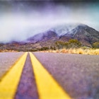 Low angle view near the yellow stripes of the road that heads towards the Franklin Mountains mountains covered by clouds, creating a misty atmosphere. Shrubs and grasses of a vivid golden yellow color grow along side the road. El Paso, Texas.