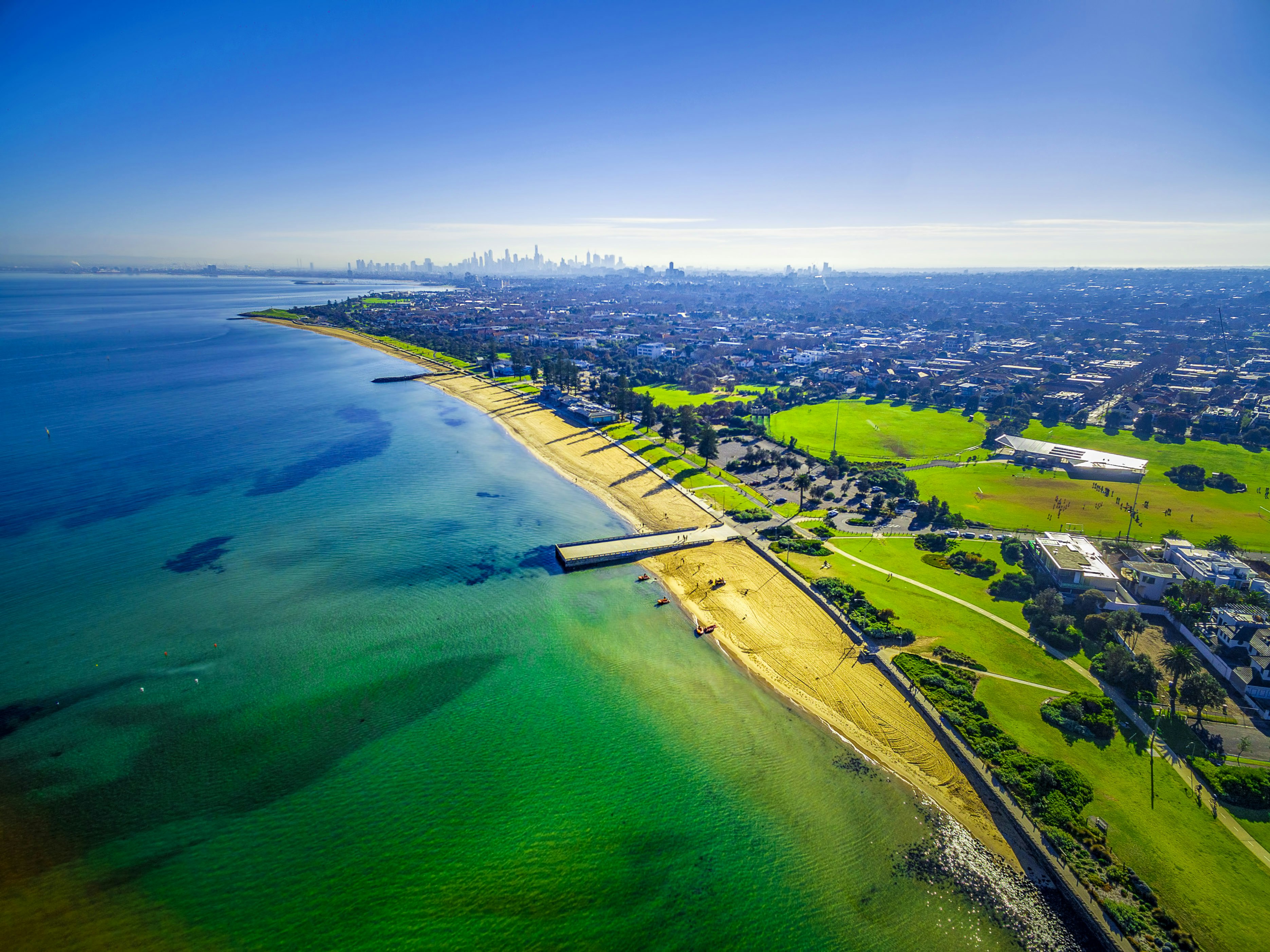 An aerial view of coastline beaches near Elwood with Melbourne CBD skyscrapers in the distance on bright sunny day.