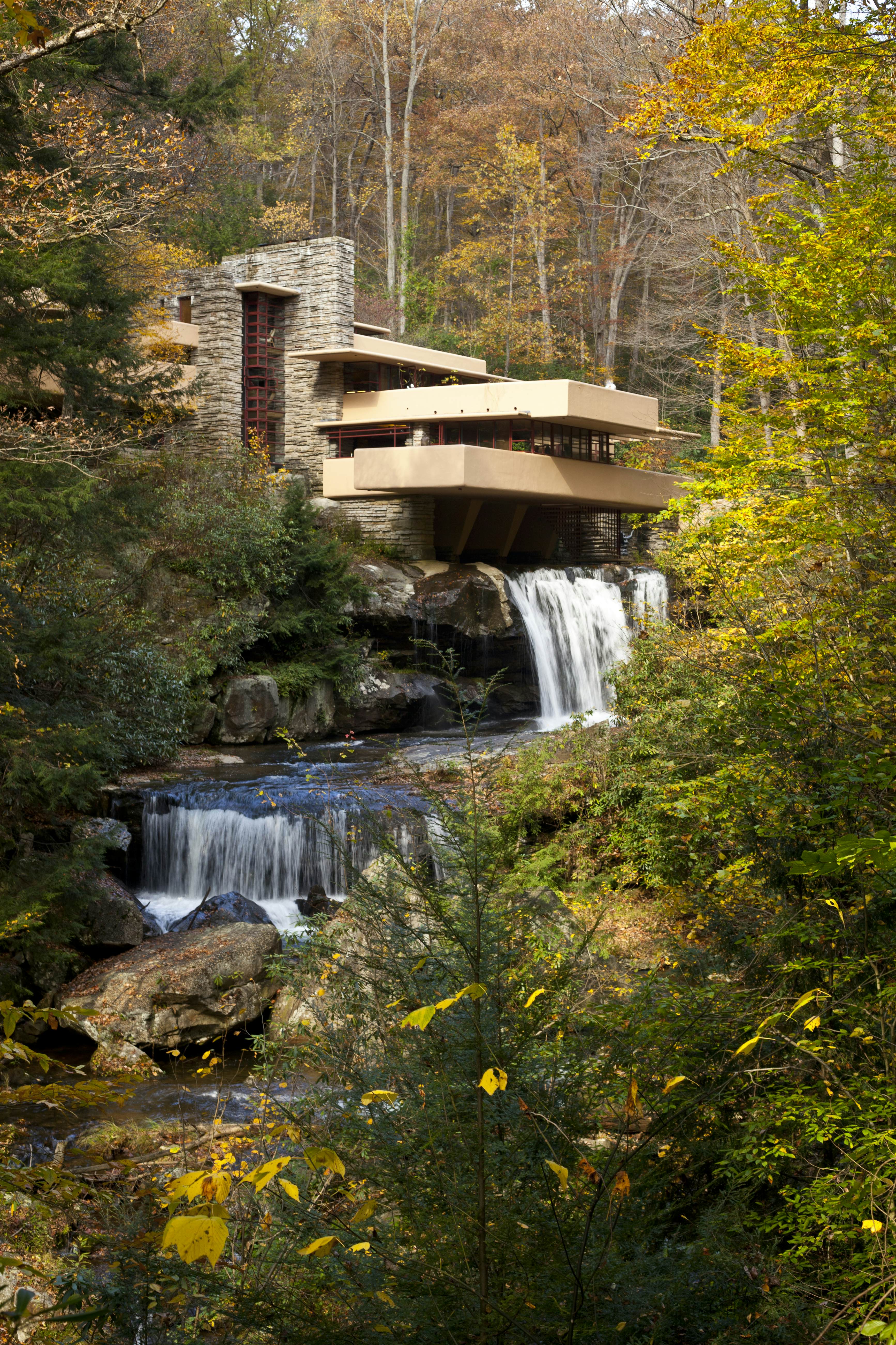 A small waterfall enclosed with tall tress and rocks, to the top right is Frank Lloyd Wright's stone structure known as Falling Water