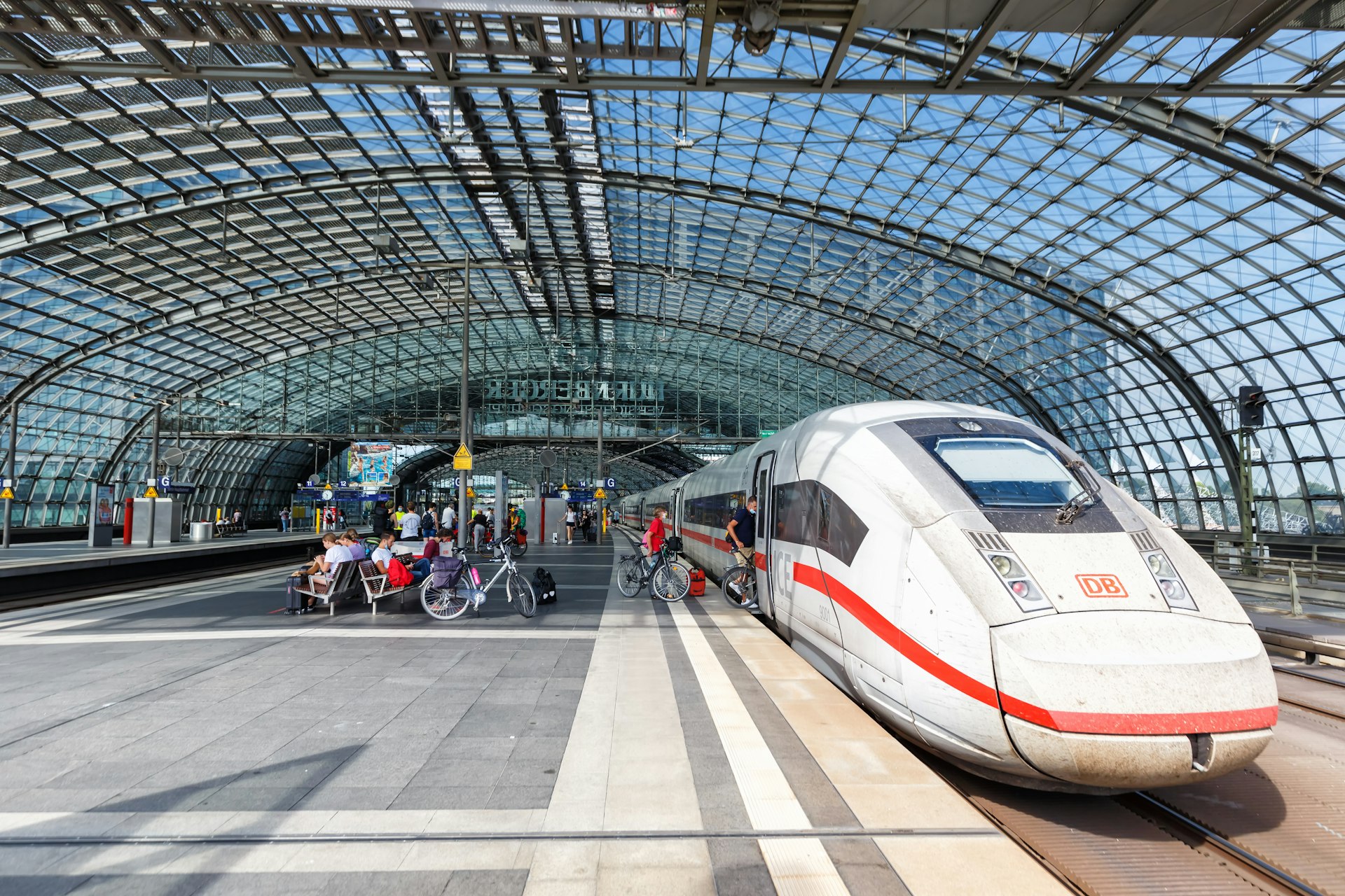 People, some with bicycles, board a high-speed train waiting at a platform in a large station with a glass roof