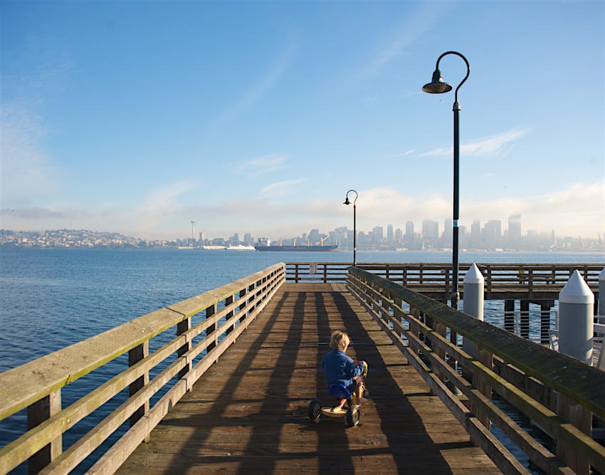 Boy rides tricycle on wooden dock looking out at Seattle skyline from Alki Beach.