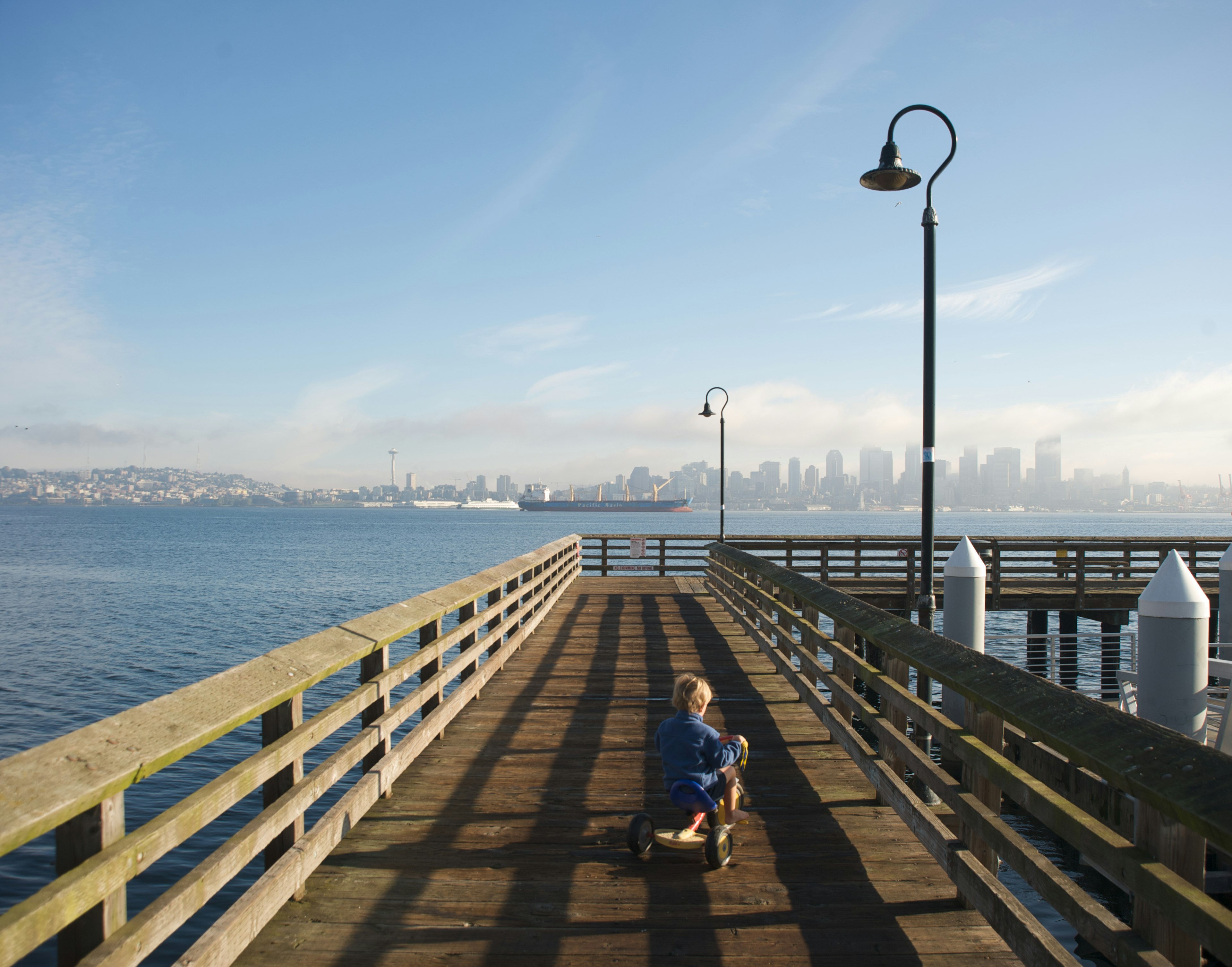 Boy rides tricycle on wooden dock looking out at Seattle skyline from Alki Beach.