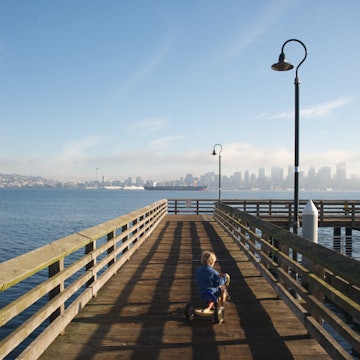 Boy rides tricycle on wooden dock looking out at Seattle skyline from Alki Beach.