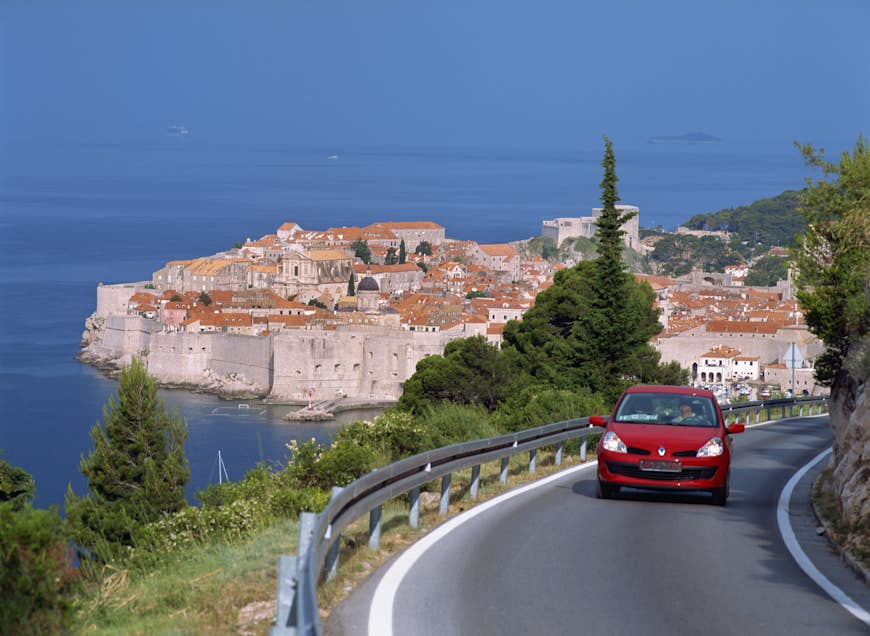 A red car coming around a curvy road with the red rooftops of Dubrovnik in the distance