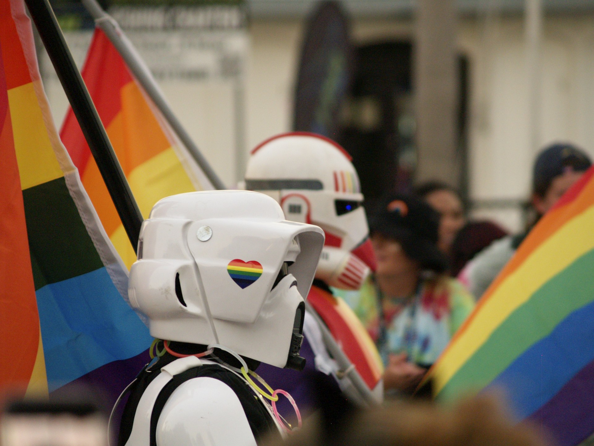A profile shot of two people in a parade wearing white Storm Trooper masks with rainbow motifs. They're carrying rainbow flags