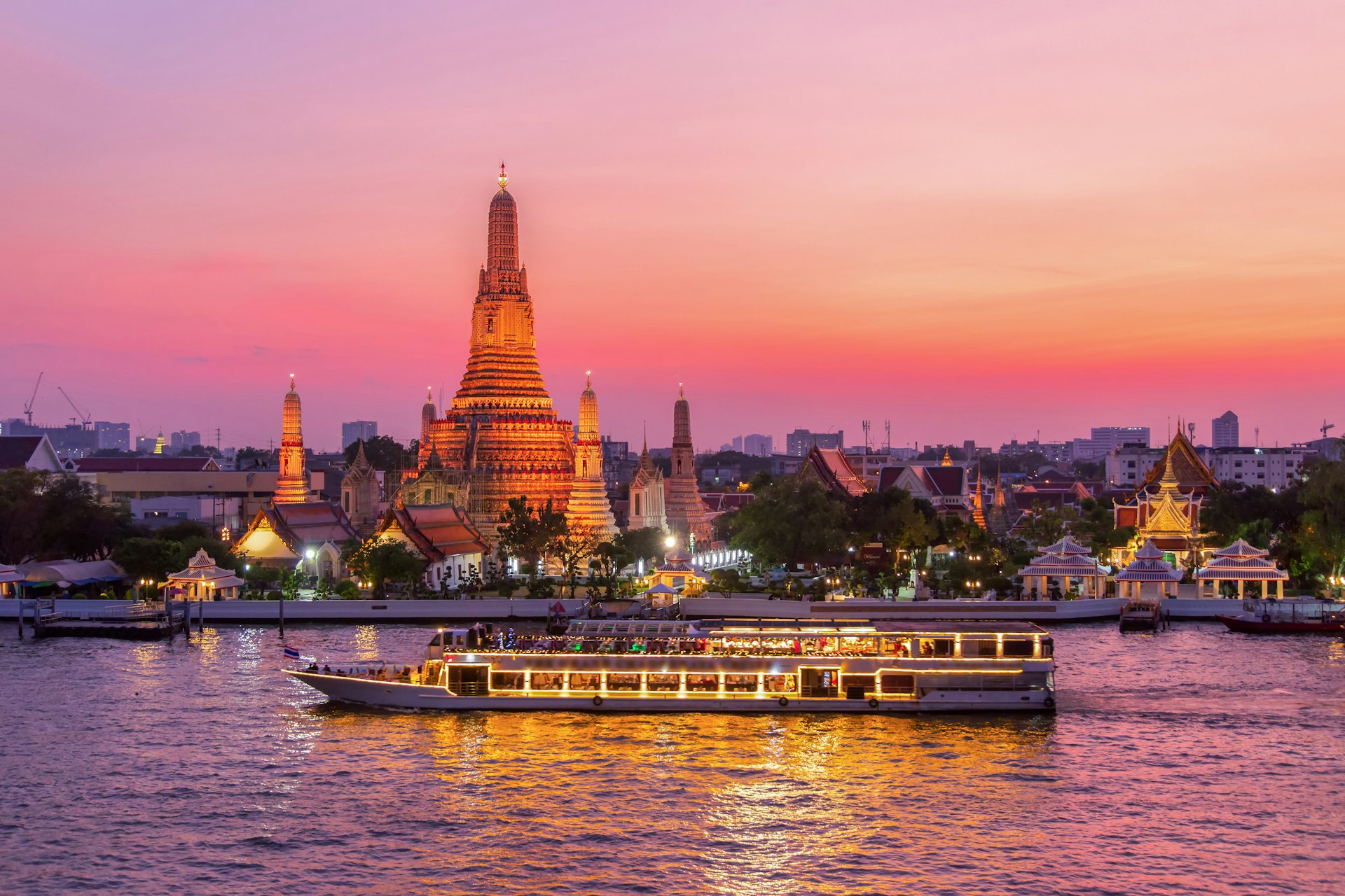 A white river cruise ships passes the stupa of Wat Arun in Bangkok under the soft glaze of an apricot-colored sunset