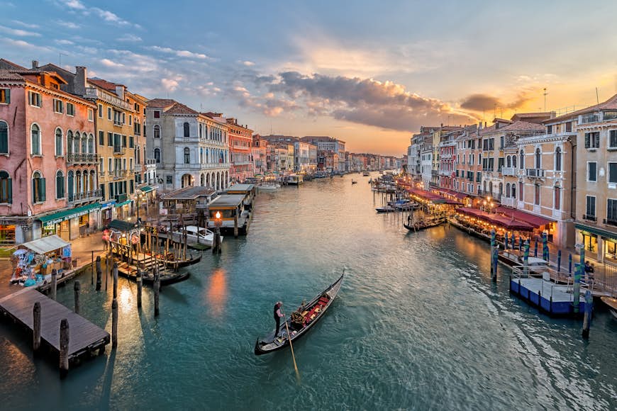 En gondol flyter nerför en bred kanal i Venedig, Italien, vid solnedgången.  Kanalen kantas av kaféer och barer där folk sitter utanför, med båtar bundna till kanalkanten.