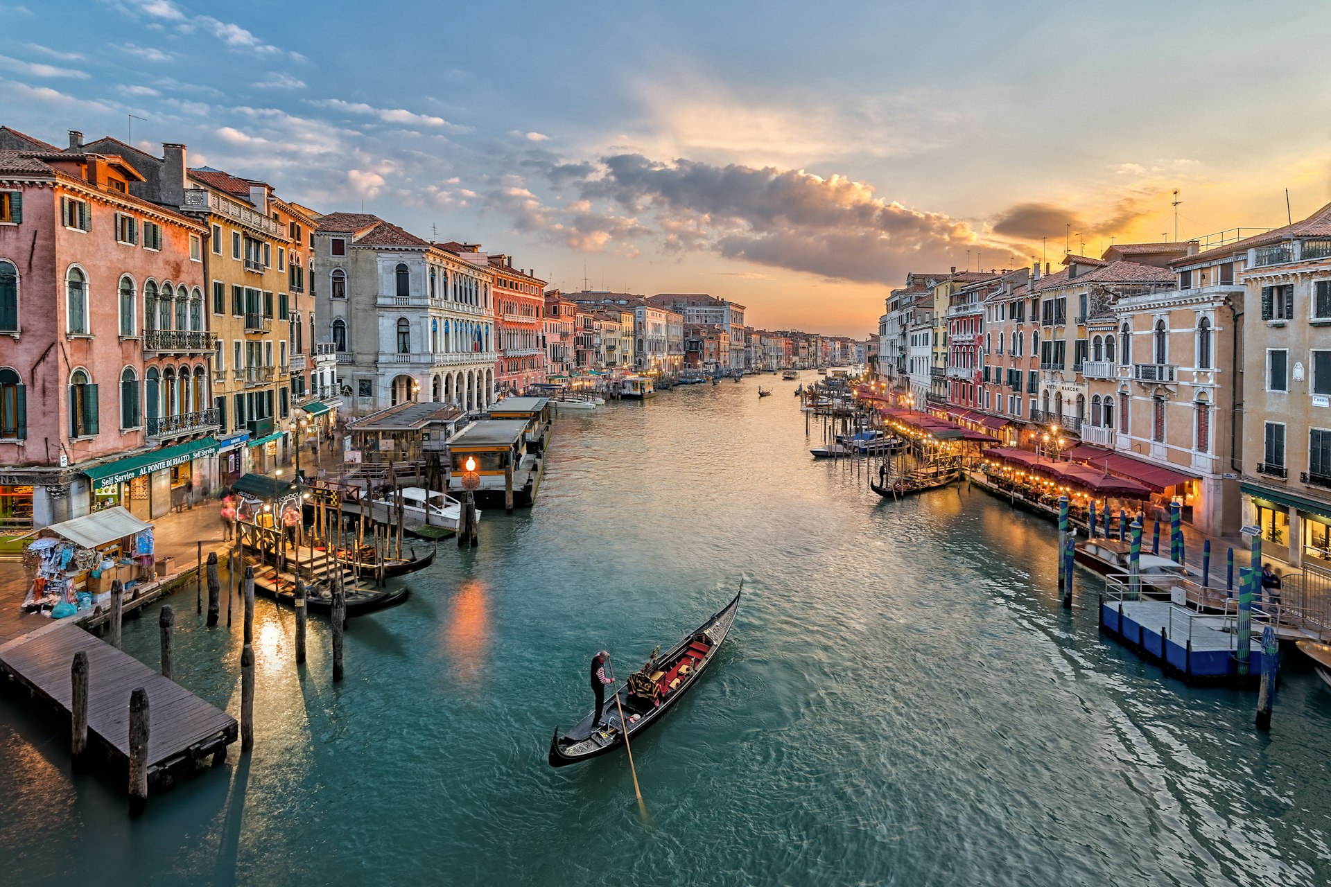 A gondola floats down a wide canal in Venice, Italy, at sunset. The canal is lined with cafes and bars where people sit outside, with boats tied up to the canal edge.