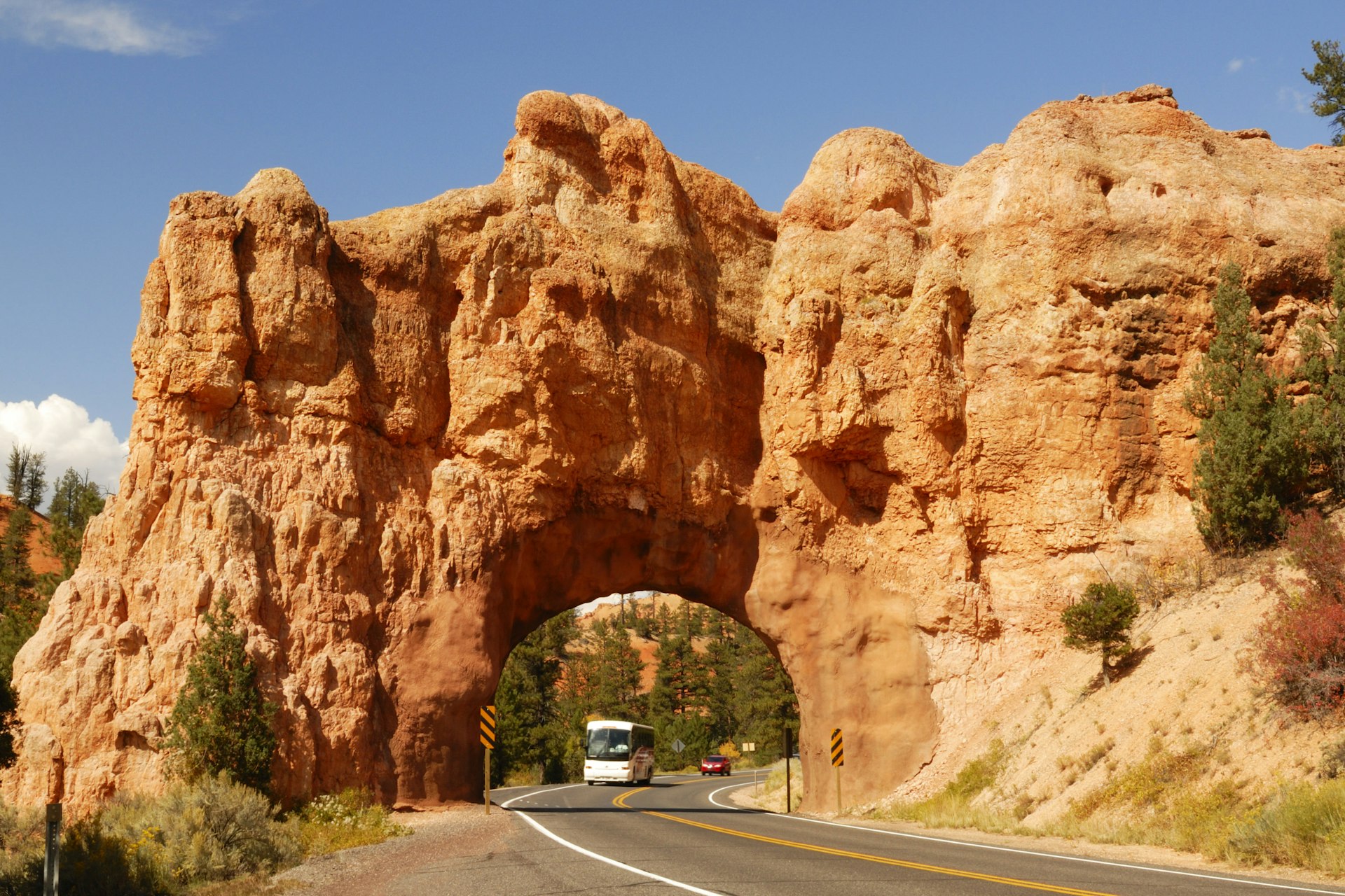 A large bus driving through a huge red rock arch that curves over a highway
