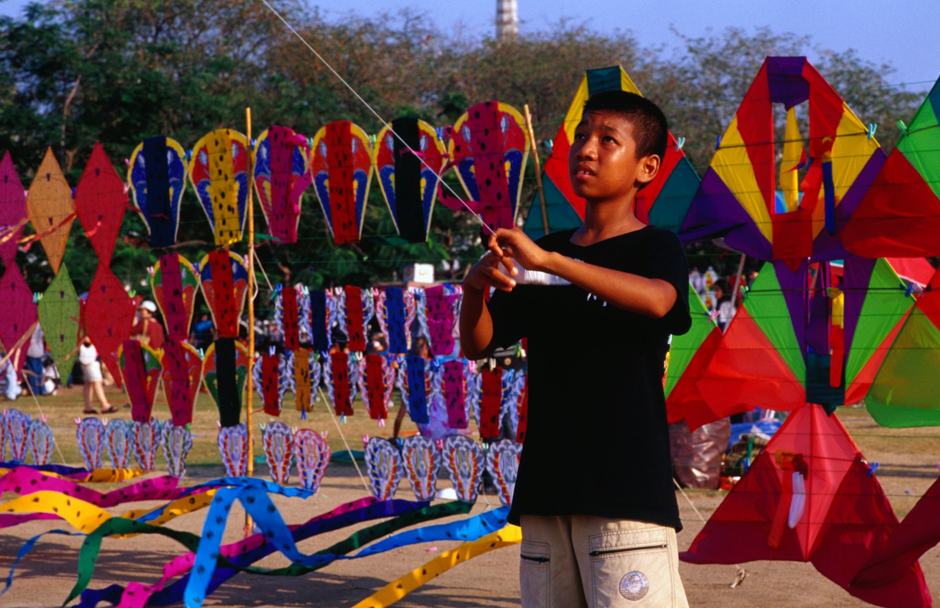 A young Thai boy flies a kite at Sanam Luang Park in Bangkok in front of a stall selling lots of colorful kites.
