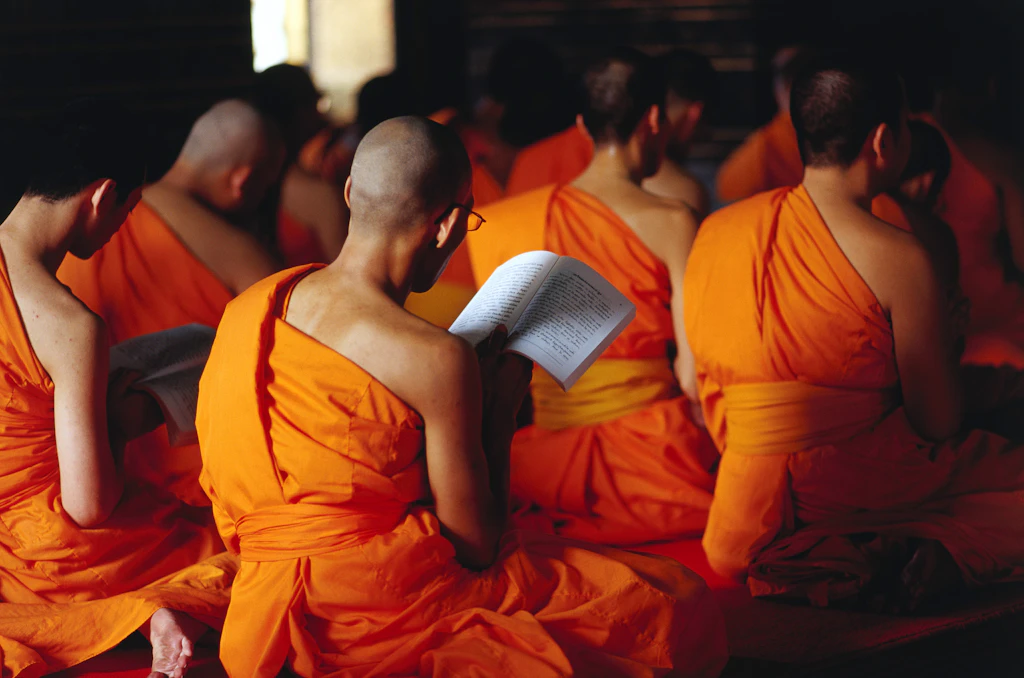 Wat Pho monks Shaved-headed monks sit cross legged at morning prayers in sermon hall at Wat Pho, some holding prayer books.