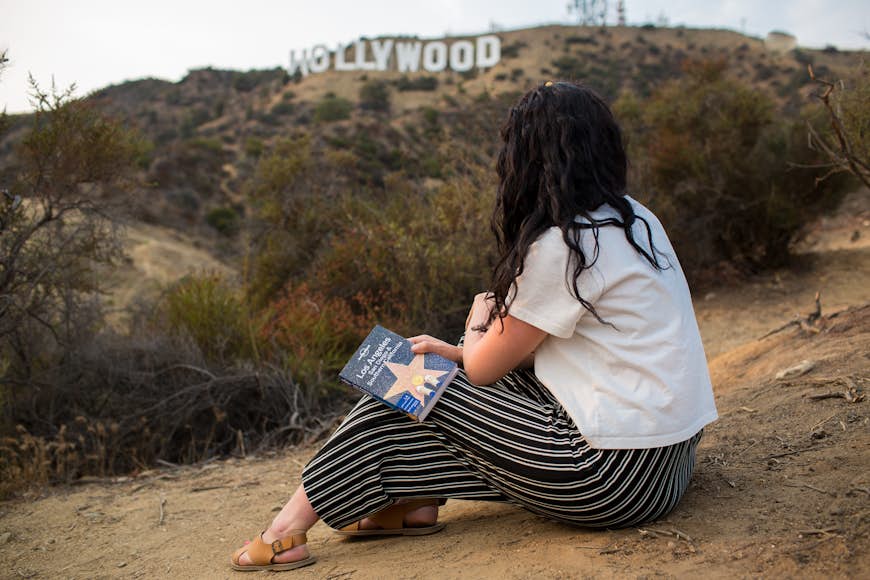Woman holding LP Los Angeles, San Diego and Southern California guidebook with Hollywood sign in background.