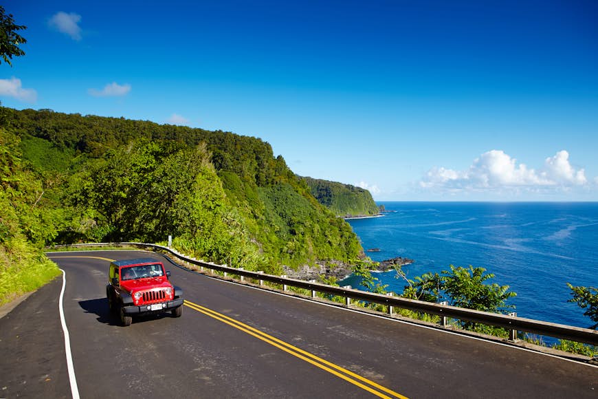 A red car follows a hilly cliff road with the sea to one side