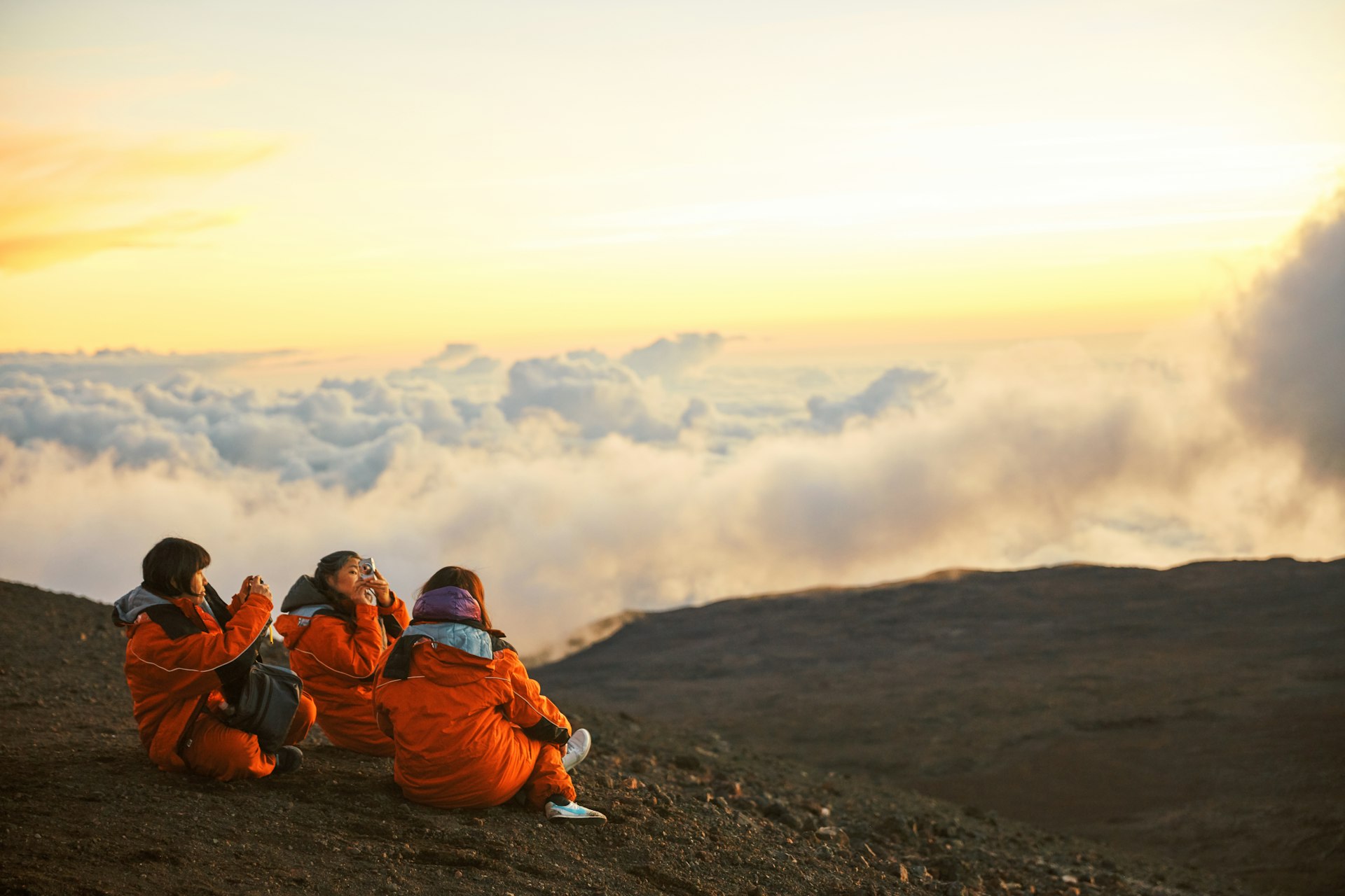 Three people sit taking photos of the sunset on a mountain above the clouds