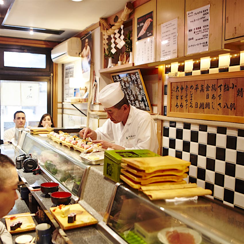 A chef carefully prepares small dishes of sushi behind a counter with two diners looking on