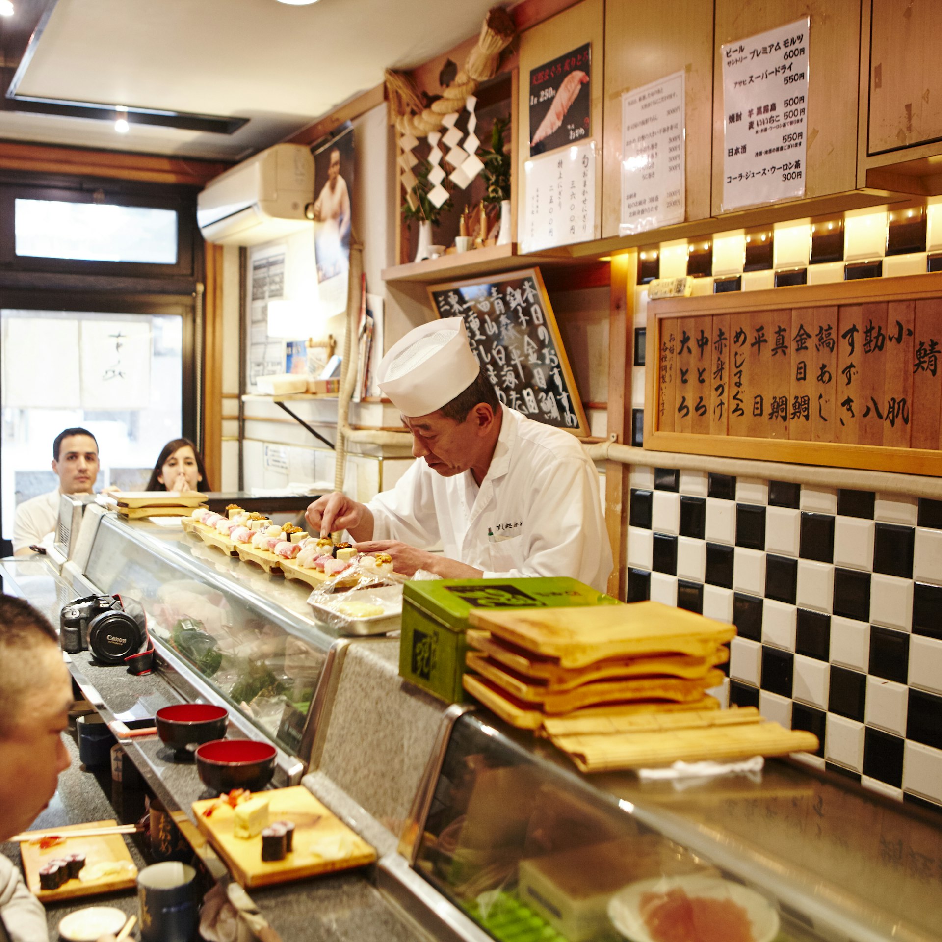 A chef carefully prepares small dishes of sushi behind a counter with two diners looking on