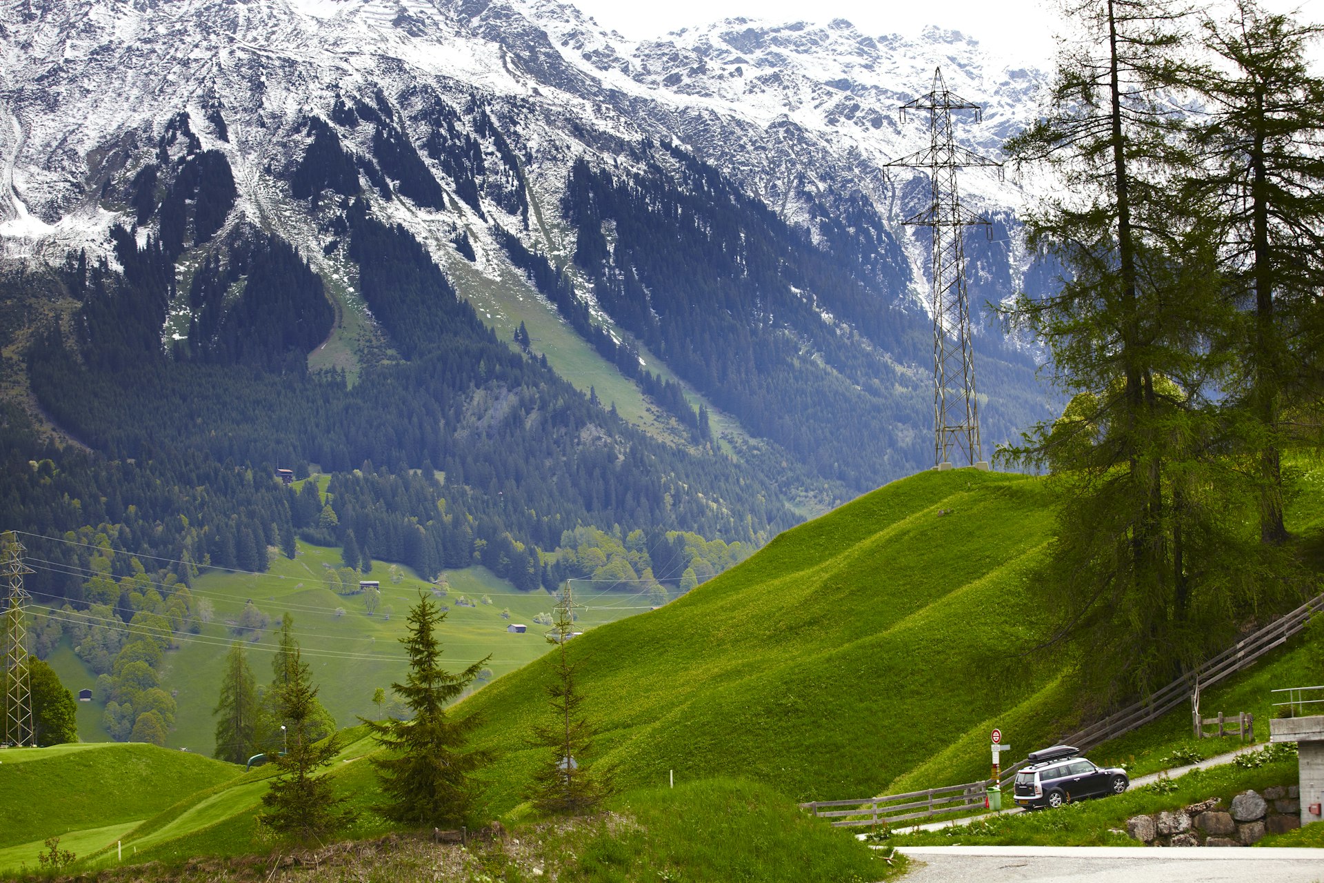 A car drives along a road through Stelvio National Park. In the background, hulking, snow-capped mountains are visible.