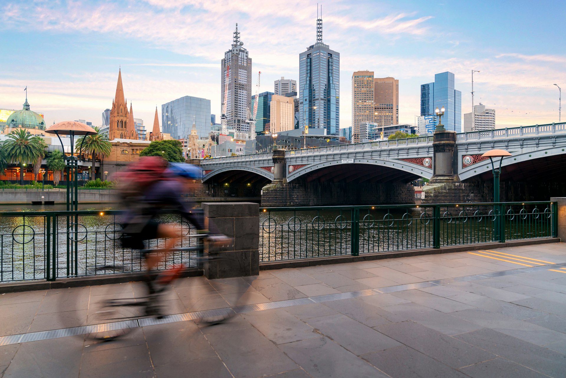 A man cycles next to the Yarra River in Melbourne, Australia. Across the river, Melbourne's main skyline is visible, with a number of towering skyscrapers.
