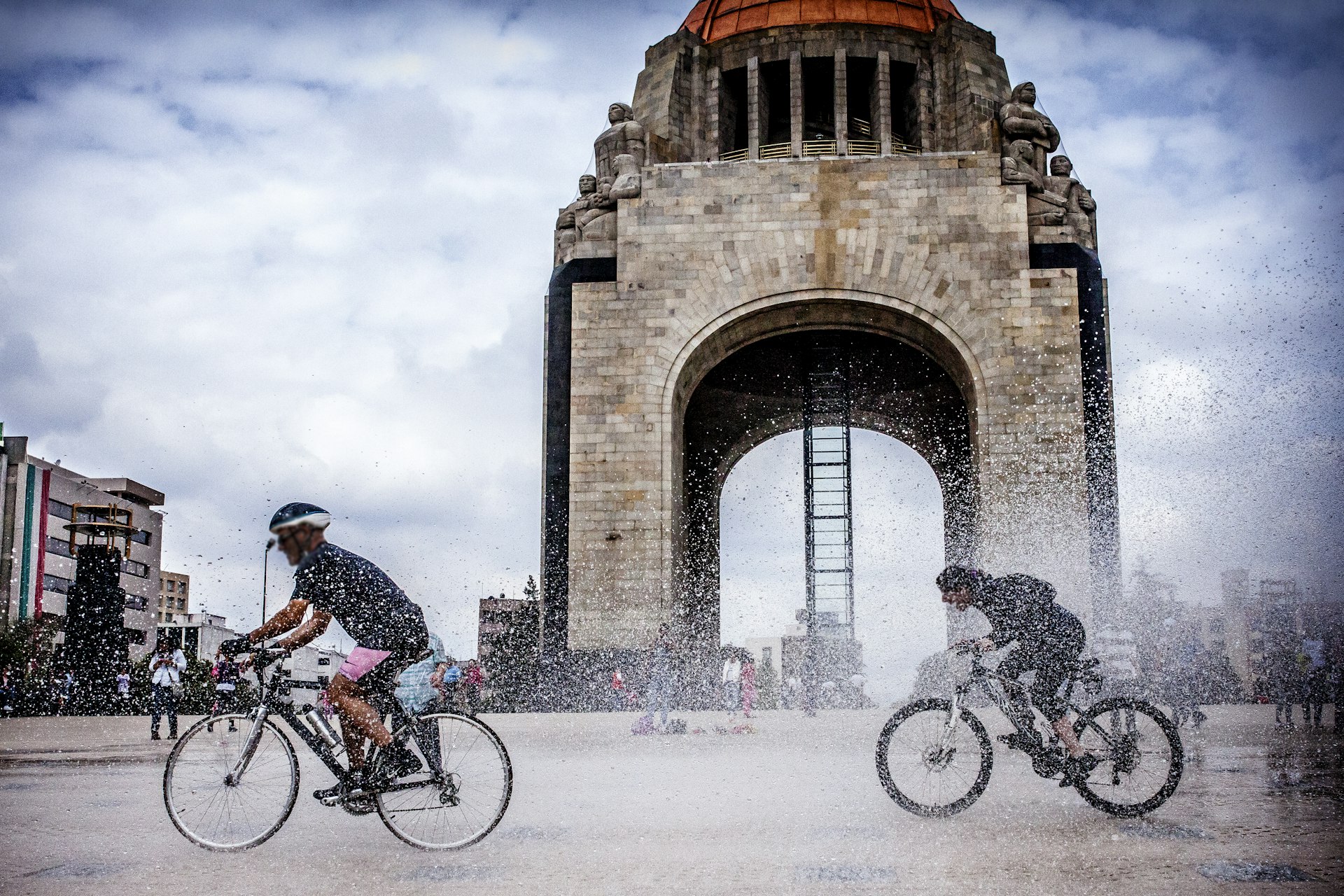 Two cyclists splash through puddles in front of a large monument
