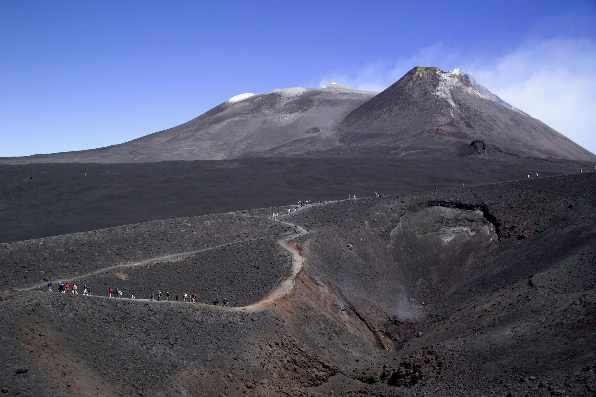 Groups of hiker walk the winding track to the blackened summit of Mount Etna in Sicily, Italy. The ash black volcano is contrasted against a clear blue sky.
