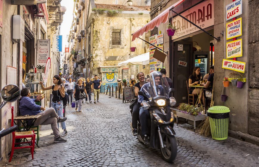 En livlig gata i Neapel.  Gatan är smal och inringad av höga hyreshus.  En motorcykel med två passagerare kör nerför gatan, medan folk äter på kaféer med bord som väller ut på trottoarerna.