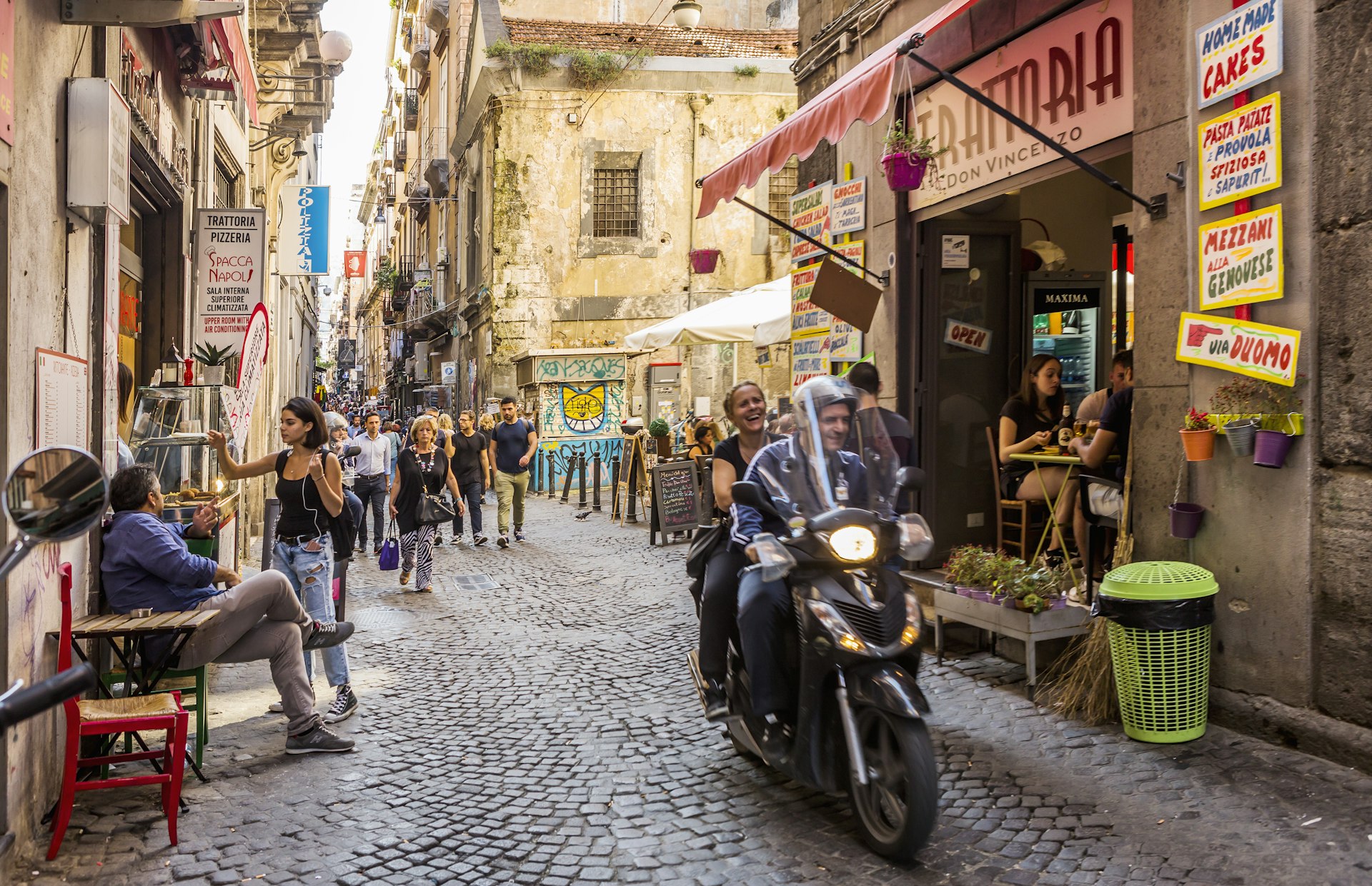 A busy street in Naples. The street is narrow and hemmed in by tall apartment buildings. A motorbike with two passengers drives down the street, while people dine in cafes with tables spilling onto the pavements.