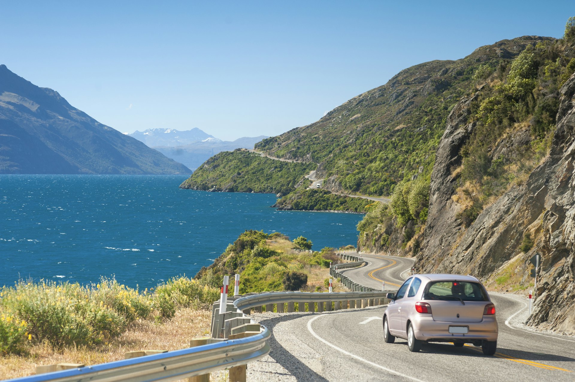 A car driving on a winding road with rocky peaks to one side and sparkling blue water to the other, on the way to Glenorchy, Queenstown, New Zealand. 