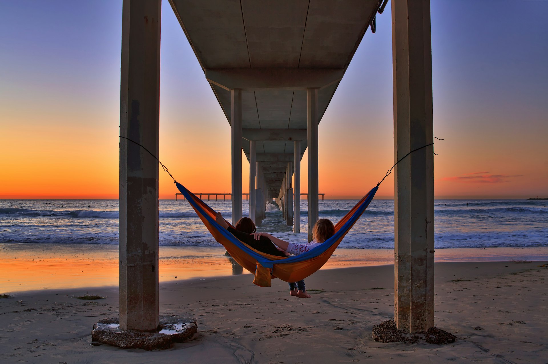 A couple relaxes in a hammock under the Ocean Beach Pier to enjoy a colorful winter sunset