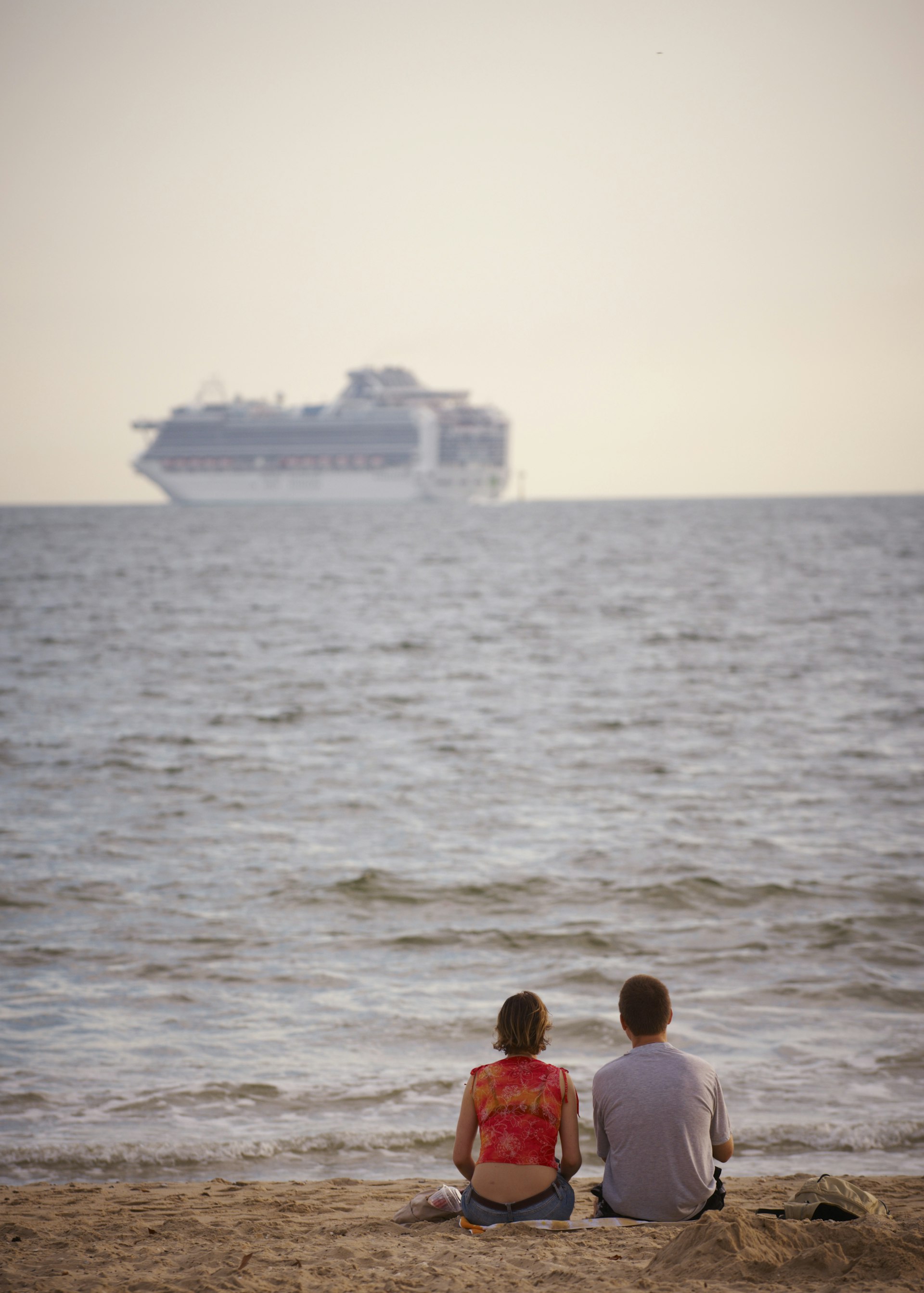 Couple sitting on sand of Port Melbourne beach watching departing cruise ship on Port Philip Bay.