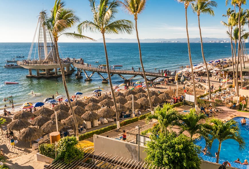 A crowded beach scene with people walking the seafront or relaxing under palm-frond shades - Puerto Vallarta Beach
