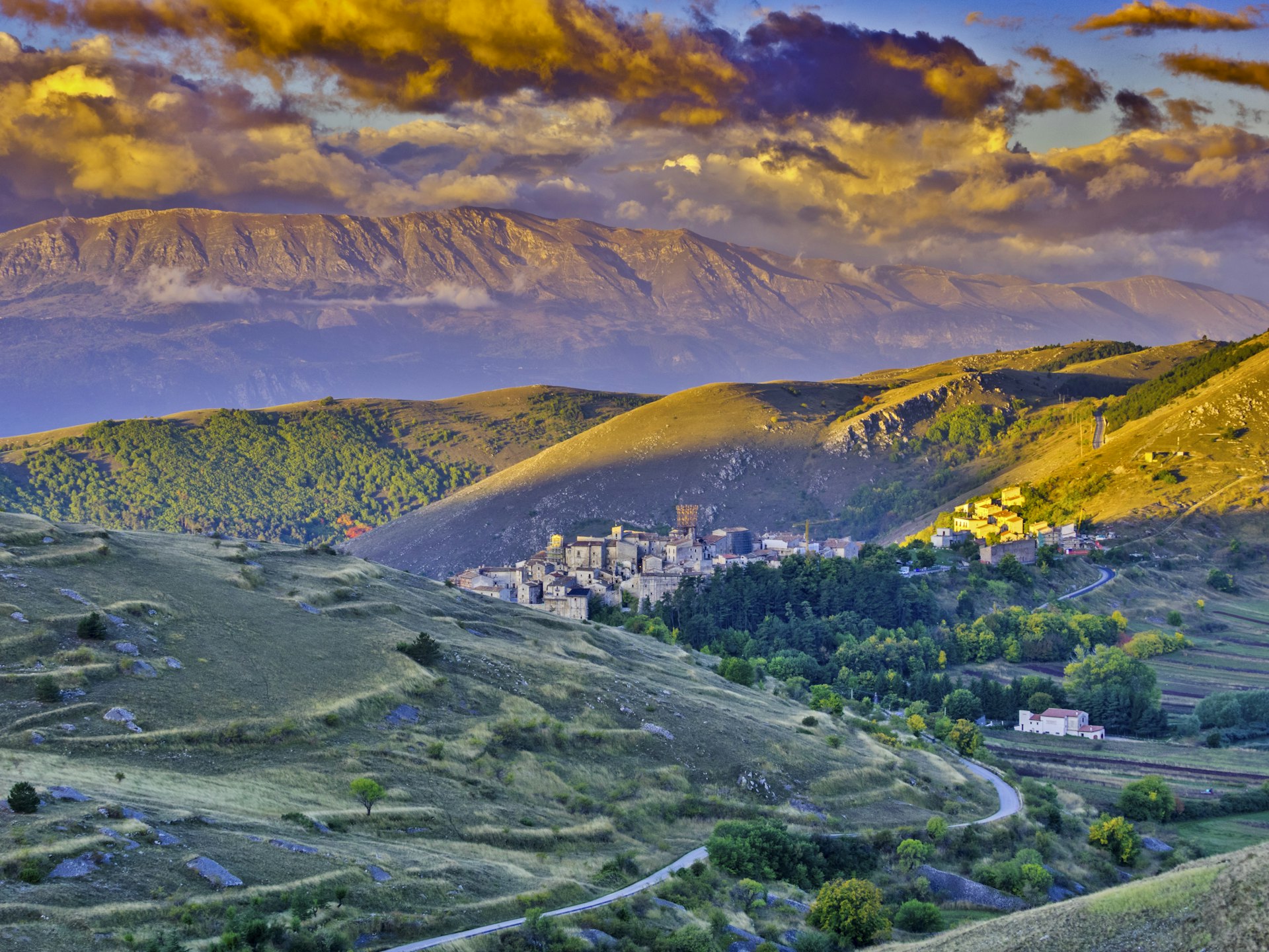 the small town of Santo Stefano di Sessanio is shown perched into a hillside in Abruzzo, Italy. All around the small town are rolling green hills, and, in the background, a set of towering mountains.