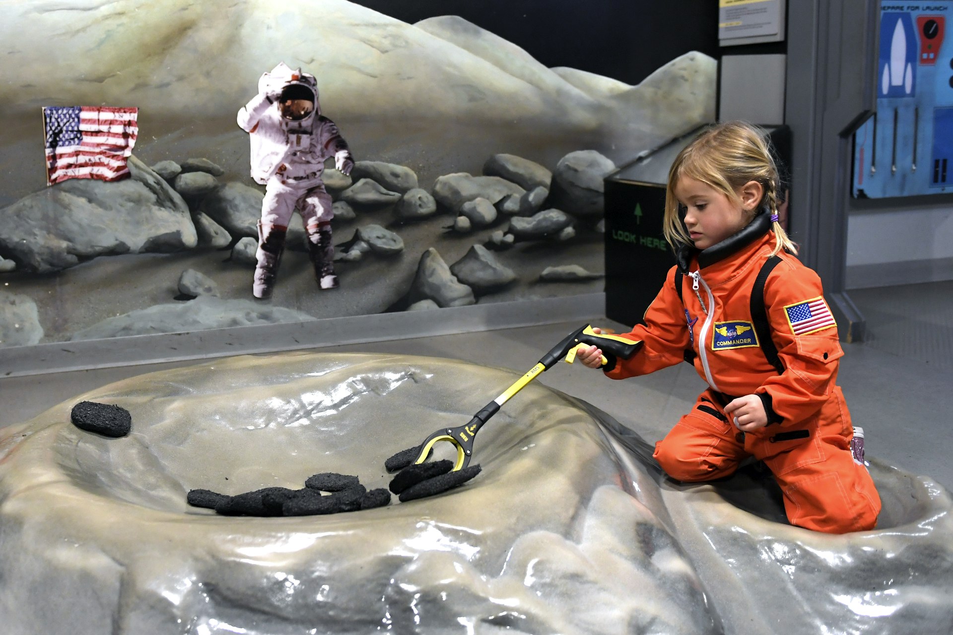 A 3-year-old girl in an orange space-style suit, picking up moon rocks at the Denver Museum of Nature & Science with her nanny Jean Wood of Denver 