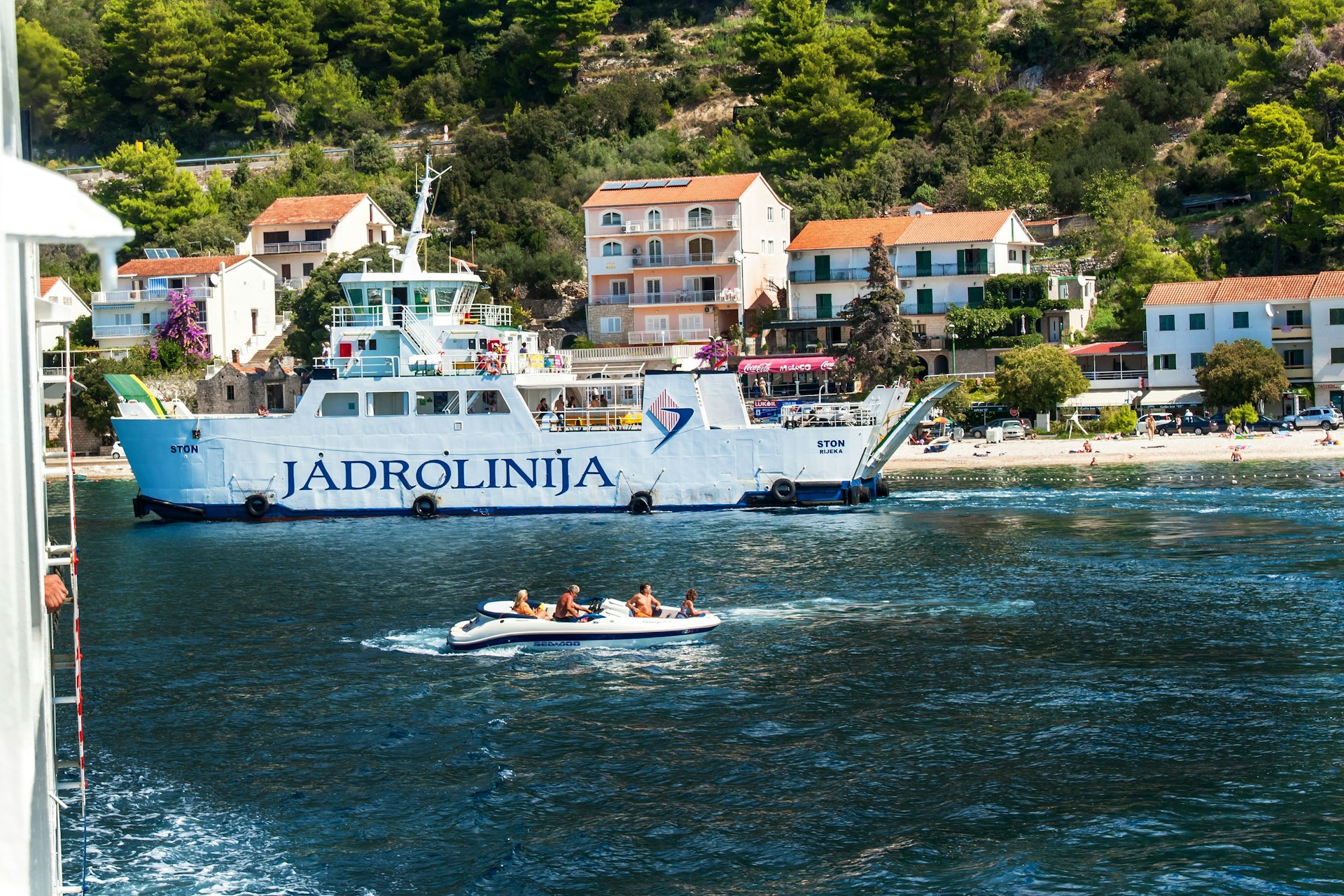 A group of kayakers pass in front of a large white car ferry with the words Jadrolinija printed on it